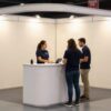 Stand modular de feria con backdrop de tela y mostrador, dos personas atendiendo a un visitante