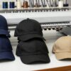 Gorras bordadas apiladas en una mesa de taller junto a una bordadora industrial y conos de hilo