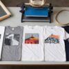 Camisetas estampadas con distintas tecnicas sobre una mesa de taller junto a un marco de serigrafia